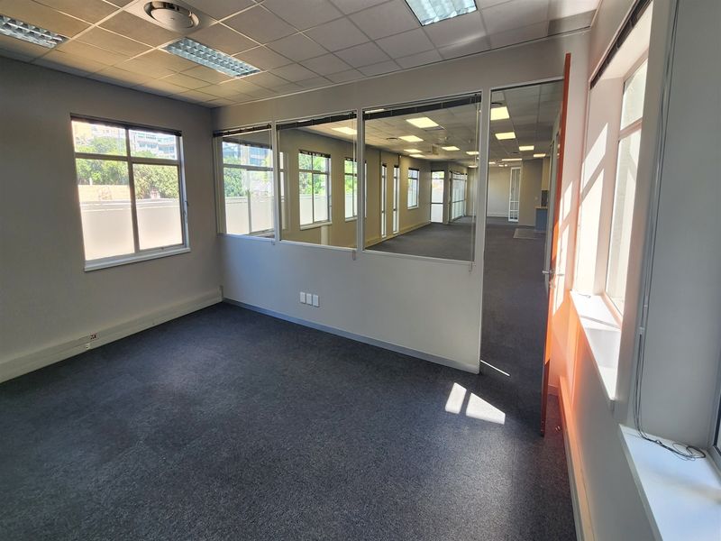 A bright, vacant office space featuring carpeted flooring, a suspended ceiling with recessed lighting, and glass partition walls providing an open, professional atmosphere.