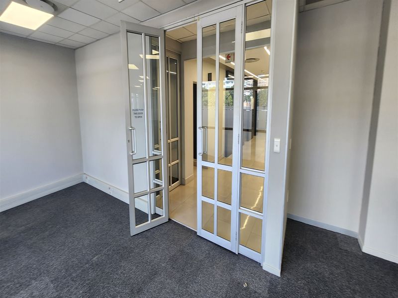 A bright, empty office space featuring glass panel double doors, carpeted flooring, and a suspended ceiling with recessed lighting.