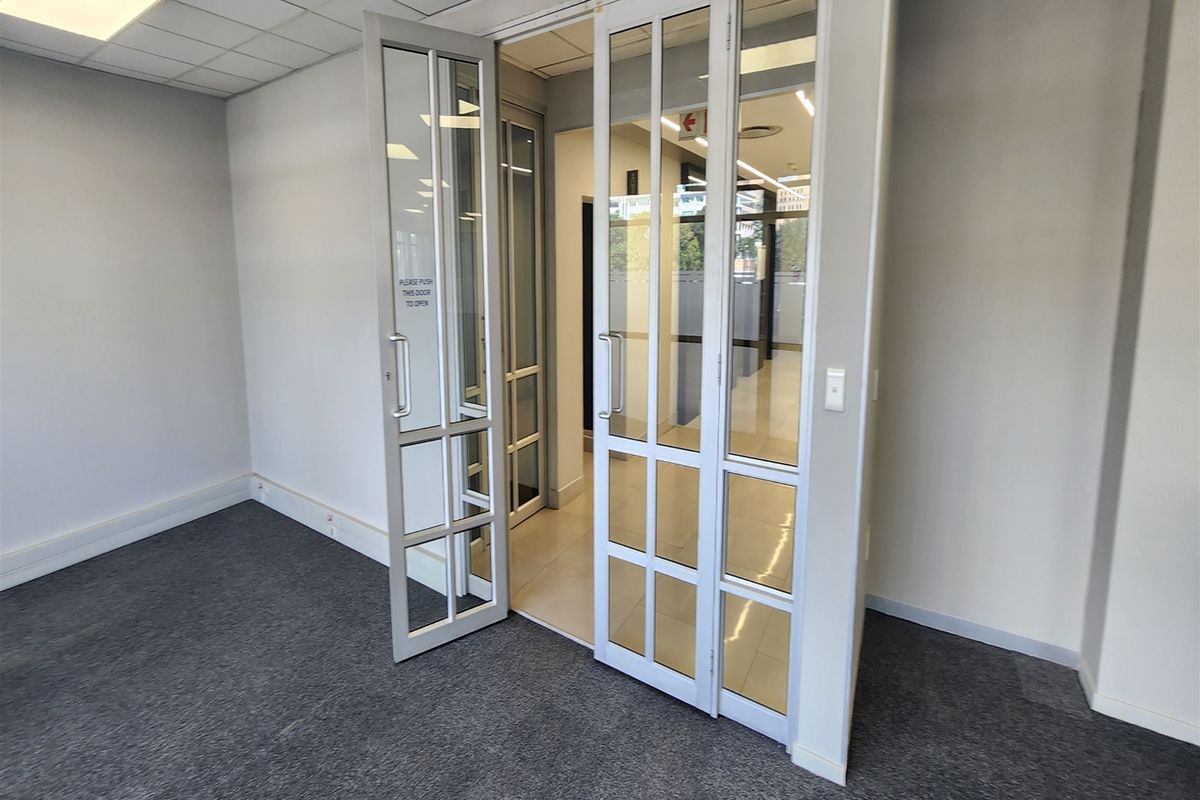 A bright, empty office space featuring glass panel double doors, carpeted flooring, and a suspended ceiling with recessed lighting.