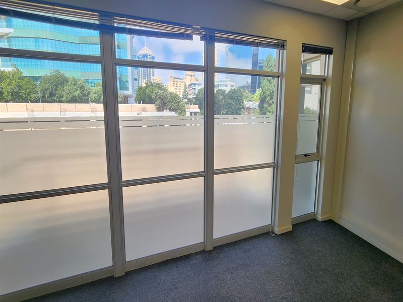 A bright, empty office space featuring large aluminium-framed windows with frosted privacy film, carpeted flooring, and a suspended ceiling.