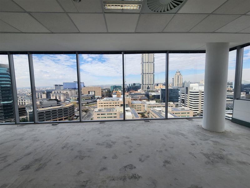 An empty, open-plan office space featuring floor-to-ceiling windows with expansive city views, a suspended ceiling, and an unfinished concrete floor.