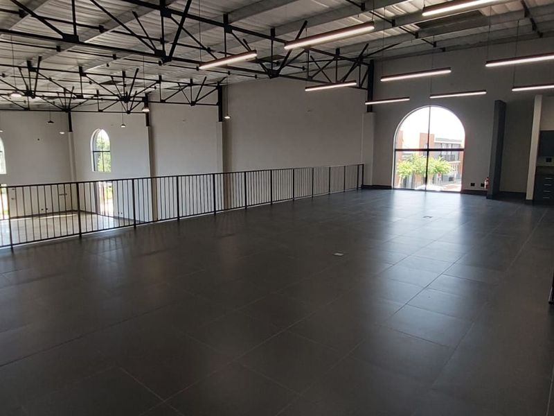 Spacious open-plan mezzanine level featuring dark tiled flooring, industrial-style exposed ceiling trusses, modern linear lighting, and arched windows overlooking the lower level.