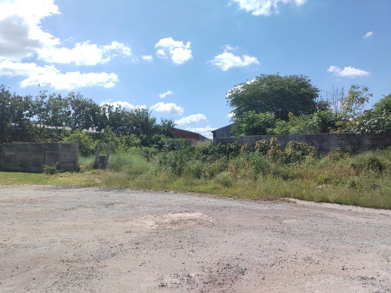 A view of a vacant commercial plot featuring an unpaved surface, overgrown vegetation, and perimeter precast concrete walling.