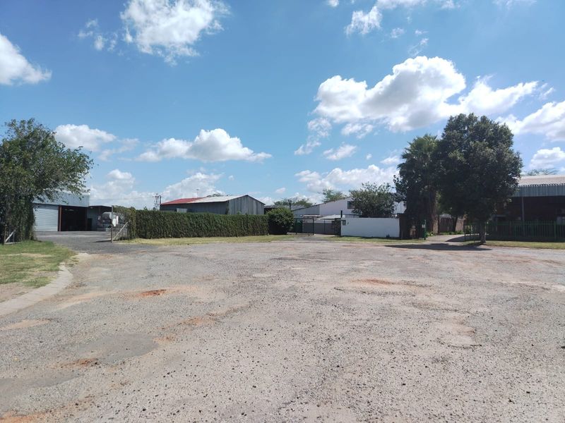 A wide-angle view of a commercial vacant land plot featuring a gravel surface, perimeter fencing, and surrounding industrial structures.