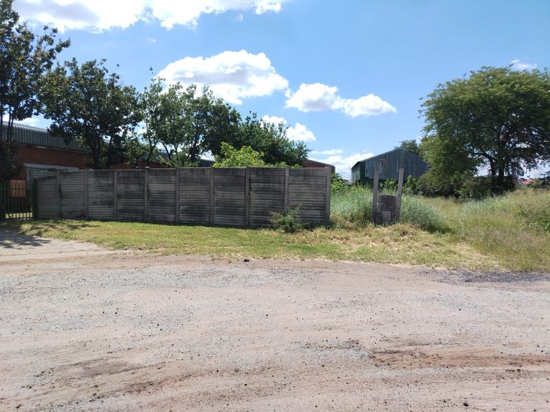 A view of a vacant commercial plot featuring a concrete boundary wall, mature trees, and overgrown grass, accessible via an unpaved road.