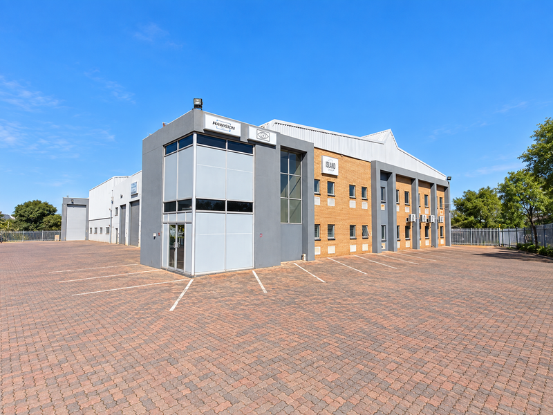 A well-maintained industrial property featuring a paved parking area, modern facade, roller shutter doors, and external air conditioning units.