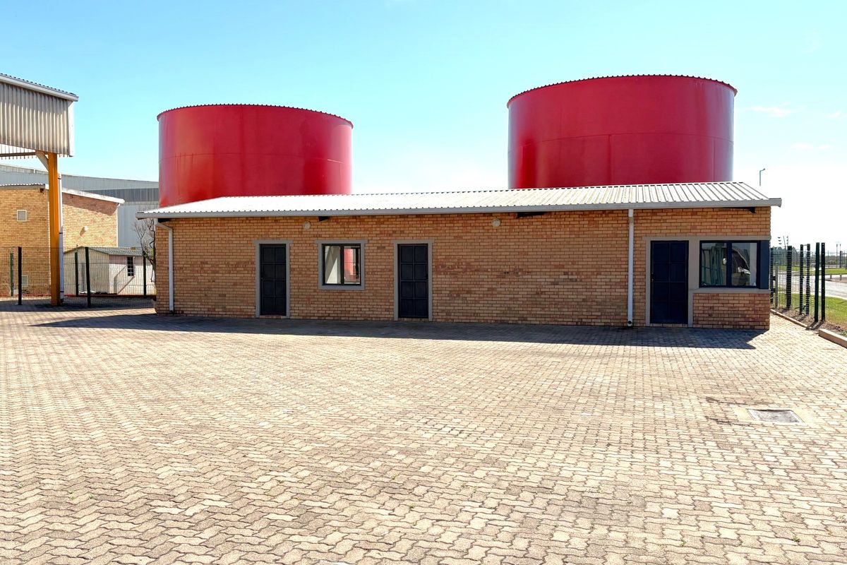 Industrial property exterior featuring a brick building with metal roofing, large red storage tanks, and a paved yard area.