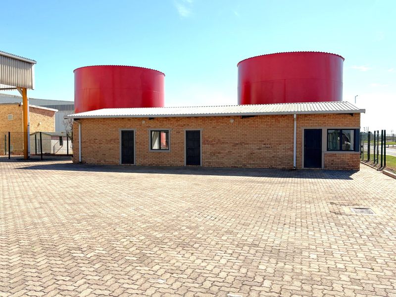 Industrial property exterior featuring a brick building with metal roofing, large red storage tanks, and a paved yard area.