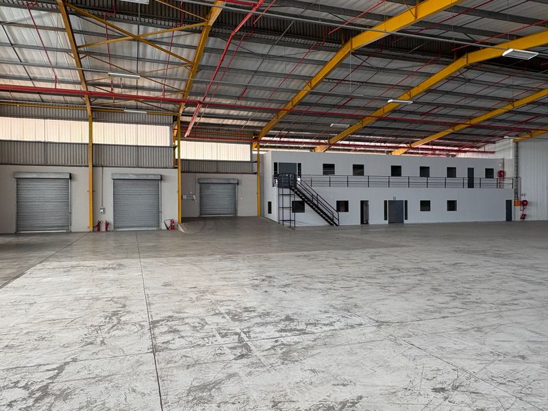 Spacious industrial warehouse featuring high ceilings with exposed steel trusses, multiple roller shutter doors, and a built-in two-level office or mezzanine structure.