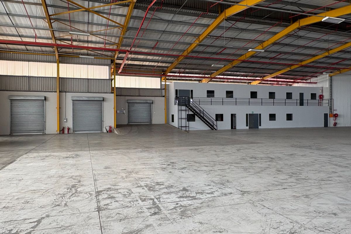 Spacious industrial warehouse featuring high ceilings with exposed steel trusses, multiple roller shutter doors, and a built-in two-level office or mezzanine structure.