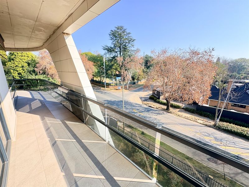 A modern, spacious balcony featuring a glass balustrade, tiled flooring, and a concrete overhang, offering an elevated view of the surrounding street and trees.