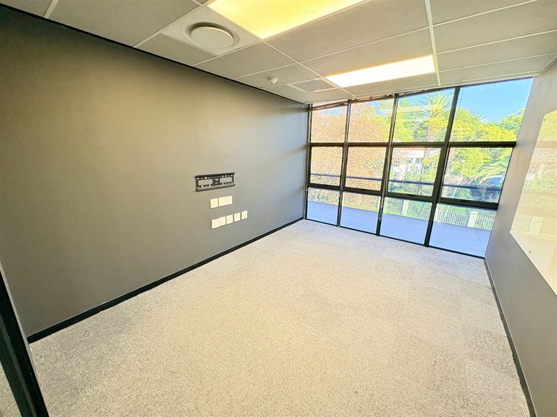 A bright, empty office space featuring large windows, carpeted flooring, a suspended ceiling with recessed lighting, and integrated wall-mounted data and power points.