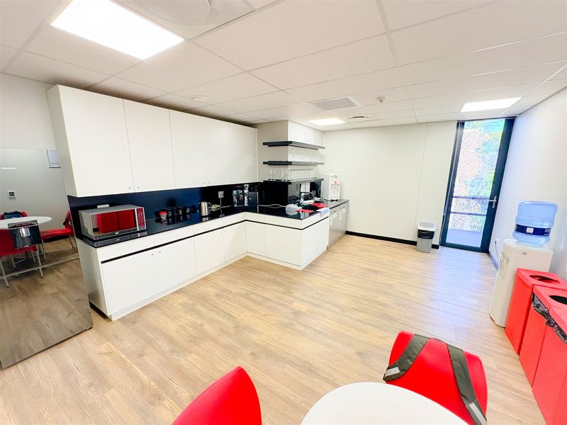 Modern office kitchenette featuring white cabinetry, black countertops, wood-look laminate flooring, and a glass door providing natural light.