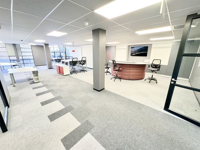 A modern, open-plan office space featuring carpeted flooring, a suspended ceiling with recessed lighting, and a central structural column.