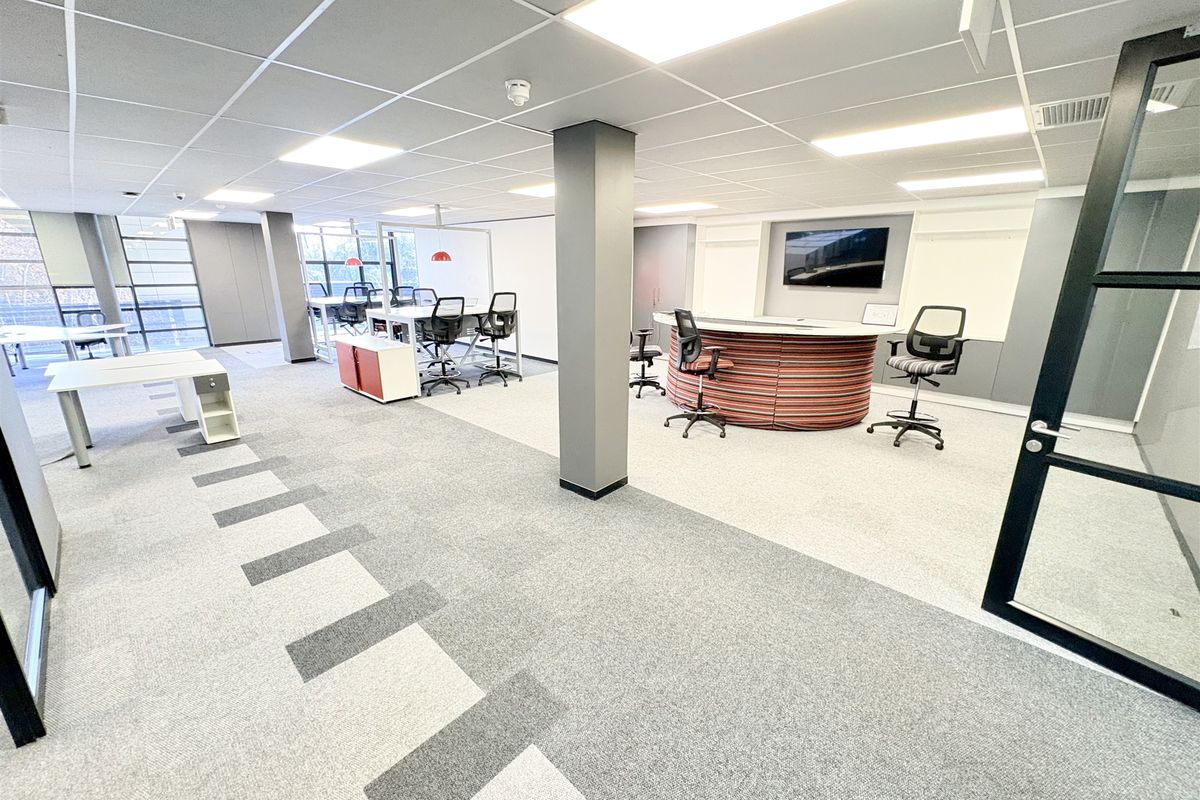 A modern, open-plan office space featuring carpeted flooring, a suspended ceiling with recessed lighting, and a central structural column.