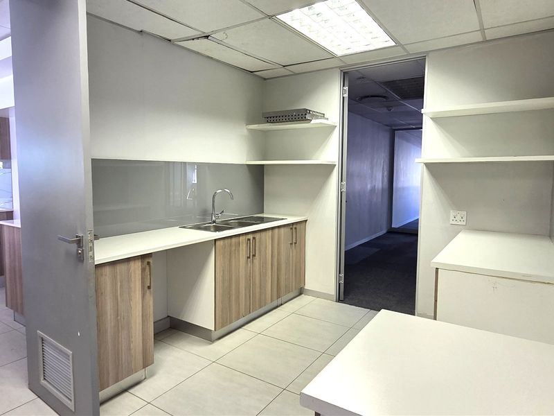 A clean, modern office kitchenette featuring wood-finish cabinetry, stainless steel sink, tiled flooring, and ample storage space with floating shelves.