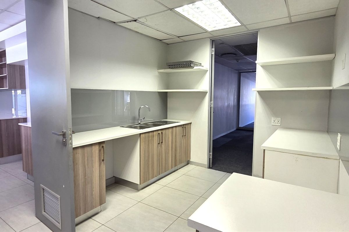 A clean, modern office kitchenette featuring wood-finish cabinetry, stainless steel sink, tiled flooring, and ample storage space with floating shelves.