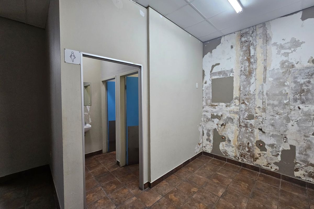 A vacant commercial restroom facility featuring tiled floors and partition walls, currently requiring significant wall repairs and cosmetic refurbishment.