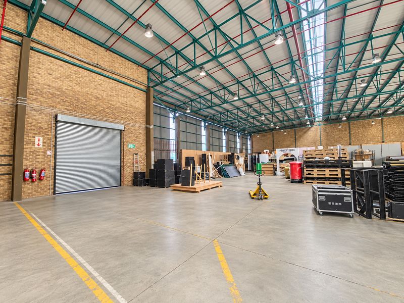 Spacious industrial warehouse unit featuring high ceilings with exposed steel trusses, concrete flooring, and a large roller shutter door for easy access.