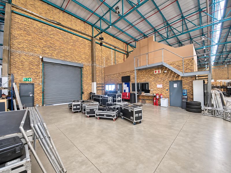 Spacious industrial warehouse unit featuring high ceilings with exposed steel trusses, a roller shutter door, concrete flooring, and an integrated mezzanine level.