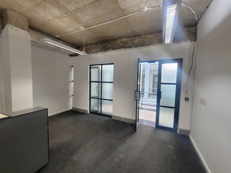 An empty office space featuring an industrial aesthetic with an exposed concrete ceiling, exposed brick accents, and modern black-framed glass doors leading to a balcony.