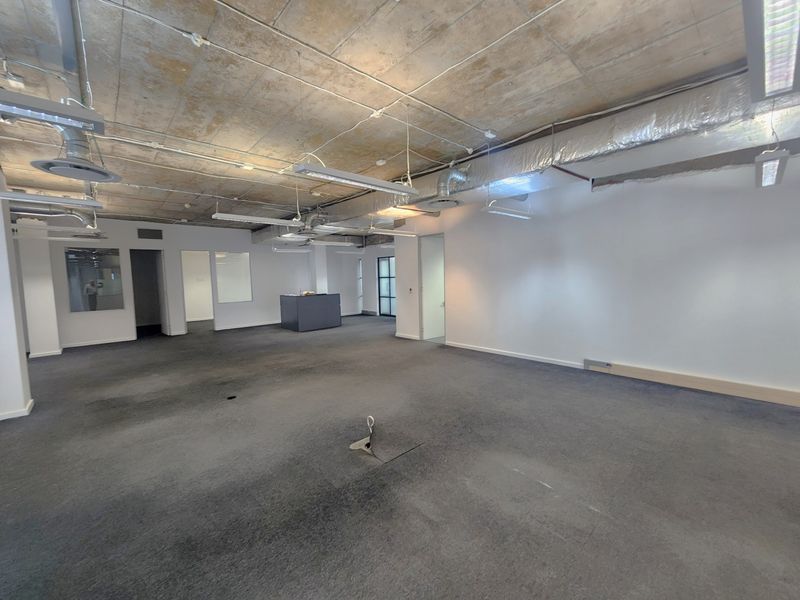 Spacious open-plan office area featuring an industrial aesthetic with exposed concrete ceilings, HVAC ducting, and neutral carpeting throughout.