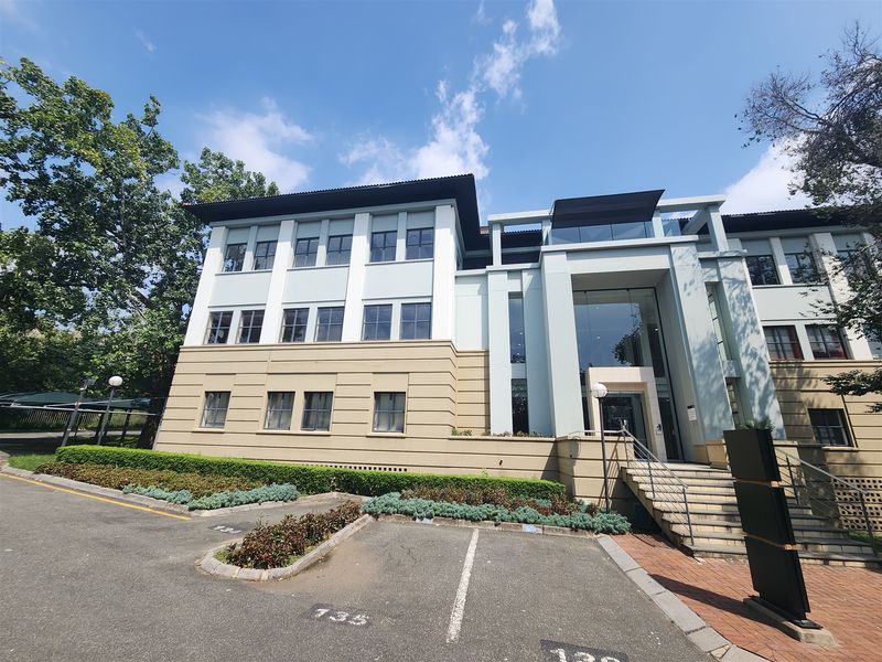 A professional multi-story office building featuring a modern glass entrance, concrete stairs, and an adjacent asphalt parking area with landscaped garden beds.