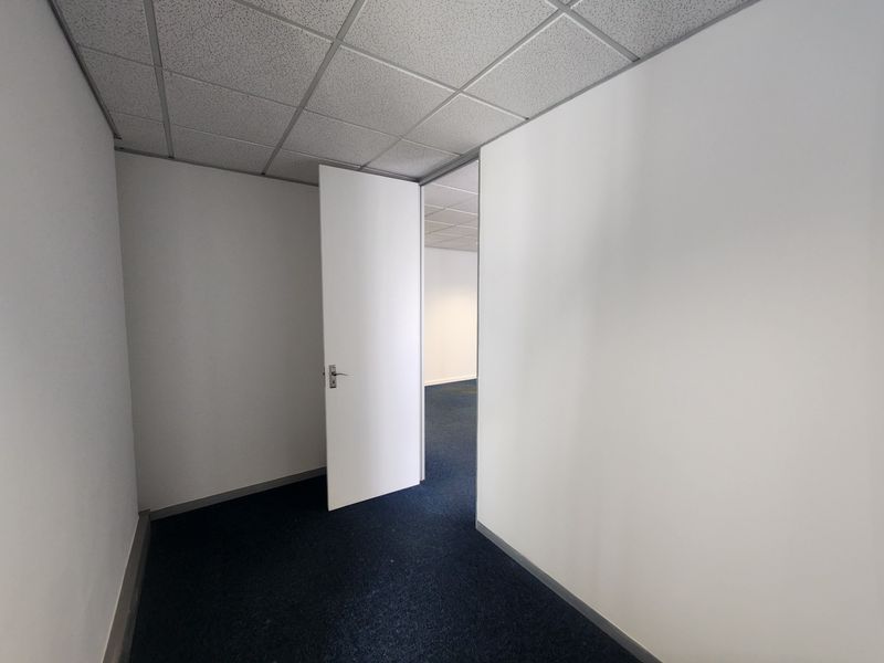 A vacant office space featuring a suspended ceiling, blue carpet flooring, and a white internal door leading to an adjacent room.