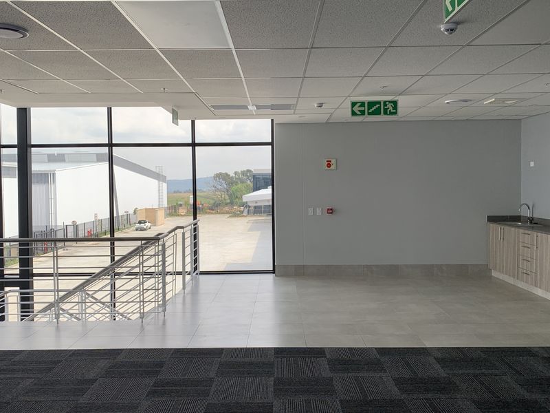 Modern open-plan office space featuring large windows, a built-in kitchenette, tiled and carpeted flooring, and industrial-style staircase access.