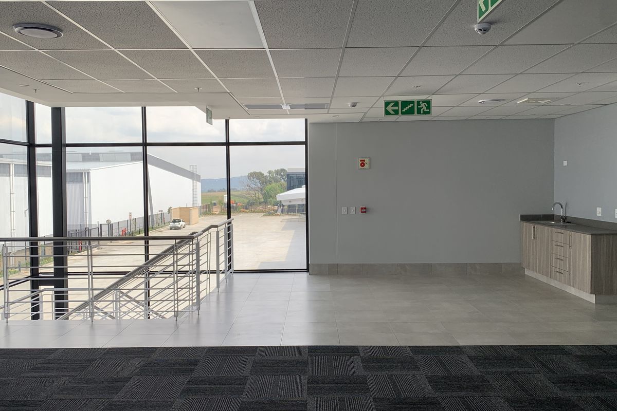 Modern open-plan office space featuring large windows, a built-in kitchenette, tiled and carpeted flooring, and industrial-style staircase access.