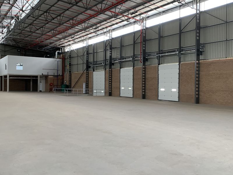 Spacious industrial warehouse featuring high ceilings, exposed steel trusses, multiple roller shutter doors, and a mezzanine level, suitable for logistics or manufacturing operations.