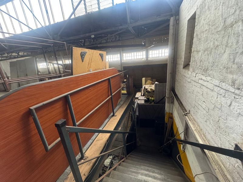 Industrial warehouse interior featuring a metal staircase leading to an upper mezzanine level with exposed brick walls and high-pitched roof structure.