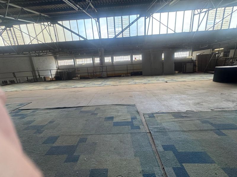 Expansive industrial warehouse space featuring high ceilings, large clerestory windows for natural light, and concrete flooring with modular carpet tiles.