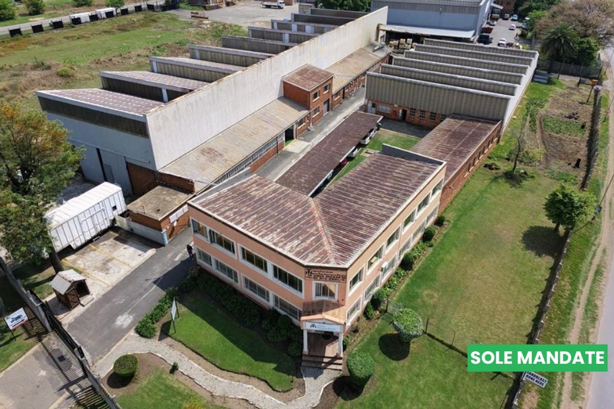 Aerial view of an industrial property featuring a large warehouse facility, attached office building, paved driveway, and surrounding landscaped grounds.