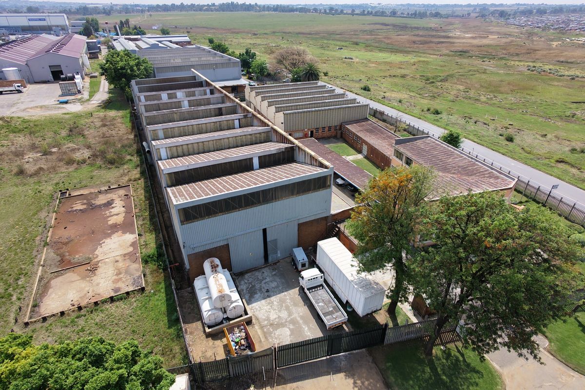 Aerial view of an industrial property featuring a large warehouse with a sawtooth roof, concrete yard, and perimeter fencing.