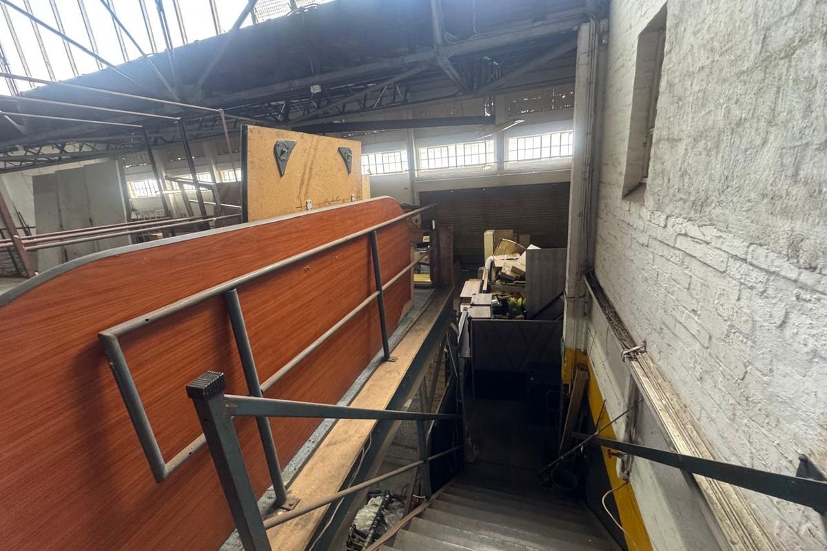 Industrial warehouse interior featuring a metal staircase leading to an upper mezzanine level with exposed brick walls and high-pitched roof structure.