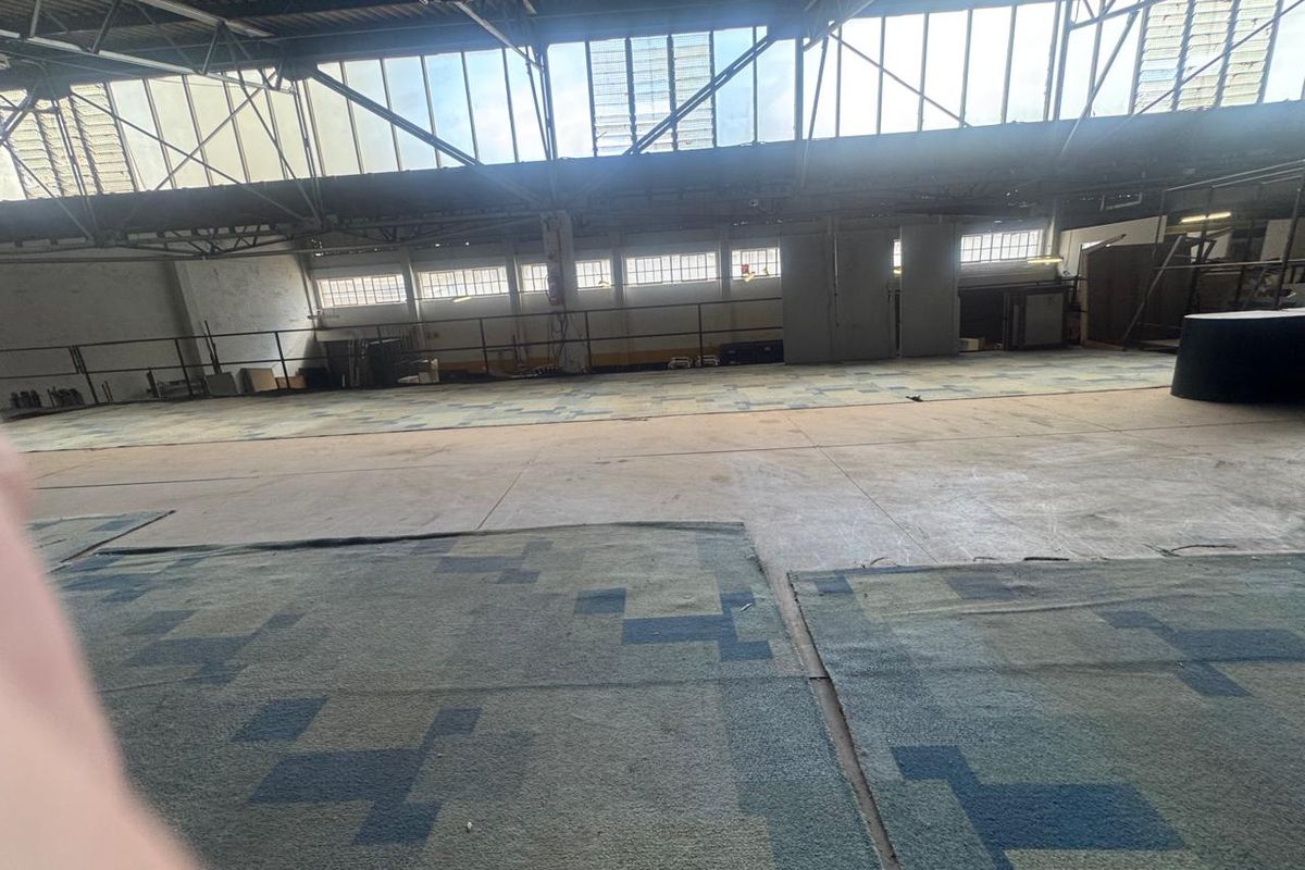 Expansive industrial warehouse space featuring high ceilings, large clerestory windows for natural light, and concrete flooring with modular carpet tiles.