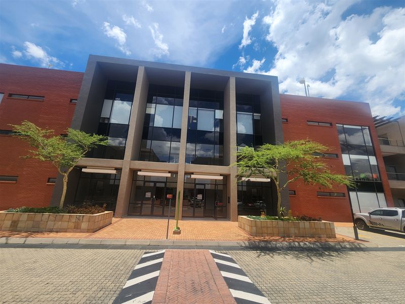 Modern office building exterior featuring a prominent brick facade, large glass curtain wall, and paved entrance area with integrated landscaping.