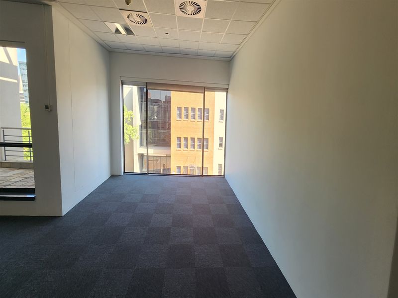 A bright, empty office space featuring carpet tiles, a suspended ceiling with recessed lighting, and a large window providing natural light.