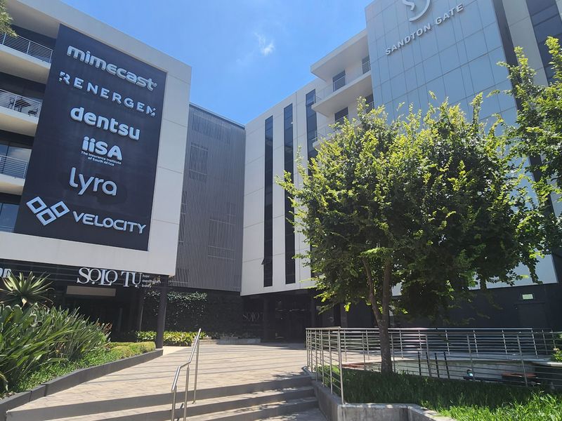 Modern commercial office building exterior featuring a paved entrance walkway, landscaped gardens, and prominent corporate signage at Sandton Gate.