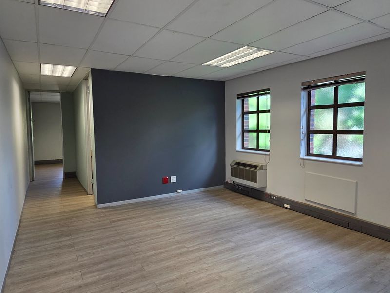 A bright, empty office space featuring laminate flooring, a suspended ceiling with recessed lighting, and a wall-mounted air conditioning unit.