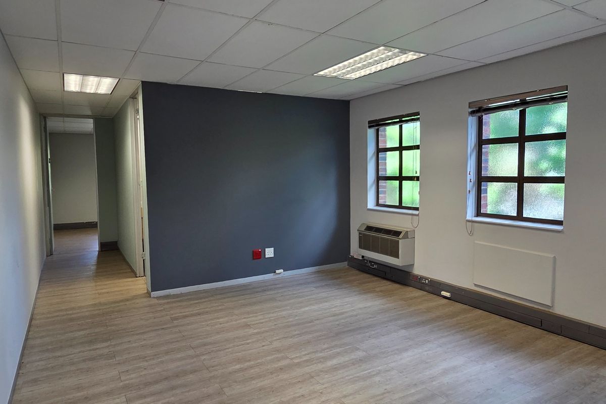 A bright, empty office space featuring laminate flooring, a suspended ceiling with recessed lighting, and a wall-mounted air conditioning unit.