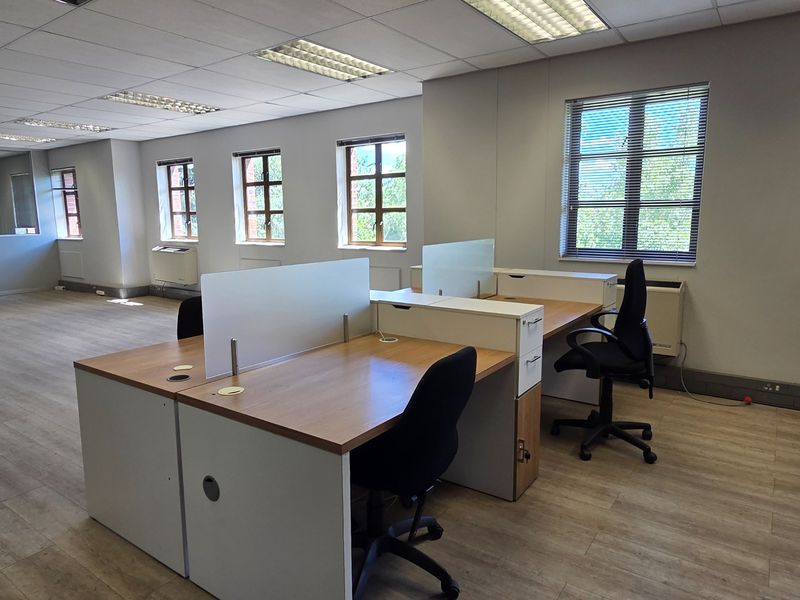 A bright, open-plan office space featuring laminate flooring, suspended ceilings with recessed lighting, and multiple windows with venetian blinds.