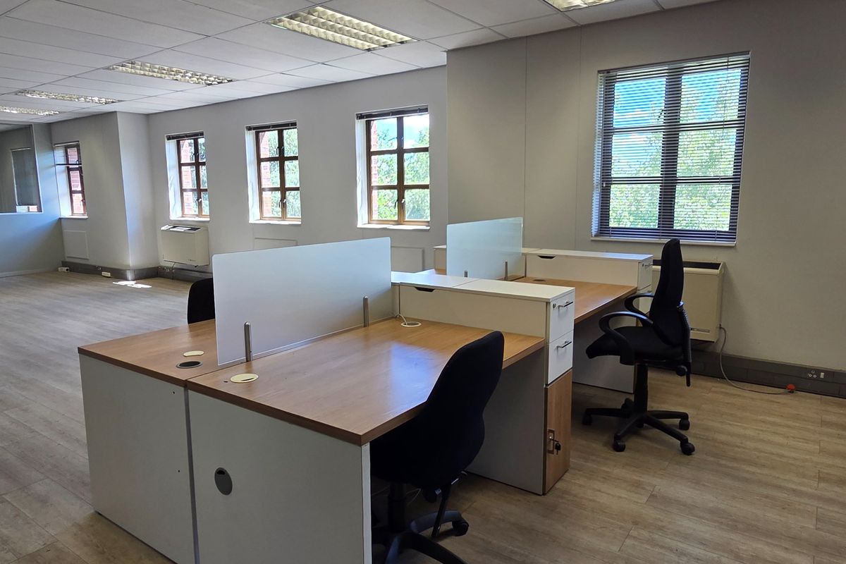 A bright, open-plan office space featuring laminate flooring, suspended ceilings with recessed lighting, and multiple windows with venetian blinds.