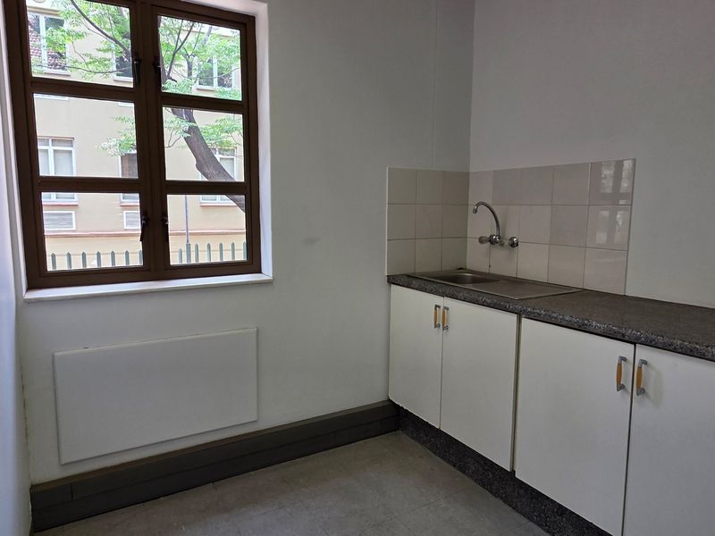 A compact, empty kitchenette featuring built-in white cabinetry, a stainless steel sink, tiled splashback, and a window providing natural light.