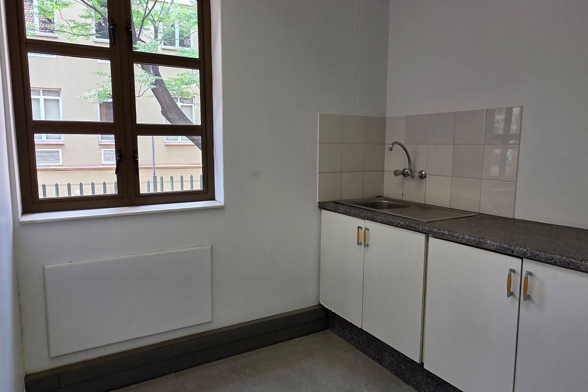 A compact, empty kitchenette featuring built-in white cabinetry, a stainless steel sink, tiled splashback, and a window providing natural light.
