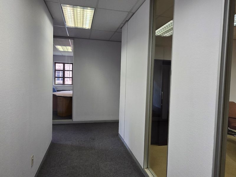A functional office hallway featuring carpeted flooring, suspended ceiling tiles with recessed lighting, and glass partition walls providing views into adjacent office spaces.