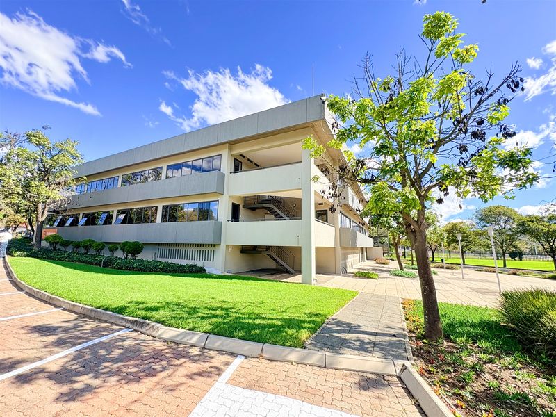 A well-maintained multi-story office building featuring a modern concrete facade, large glass windows, external staircases, and surrounding landscaped grounds with paved parking.