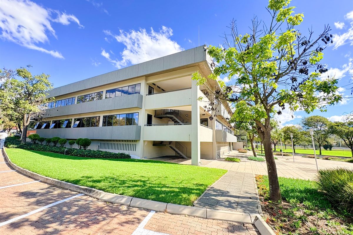 A well-maintained multi-story office building featuring a modern concrete facade, large glass windows, external staircases, and surrounding landscaped grounds with paved parking.