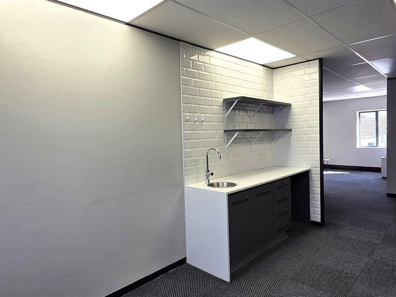 A modern office kitchenette area featuring a subway tile backsplash, built-in cabinetry with a stainless steel sink, and floating shelves in an open-plan space.